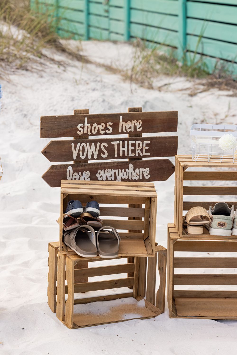 Rustic wooden sign reading shoes here vows there love everywhere at a beach wedding with crates holding sandals.