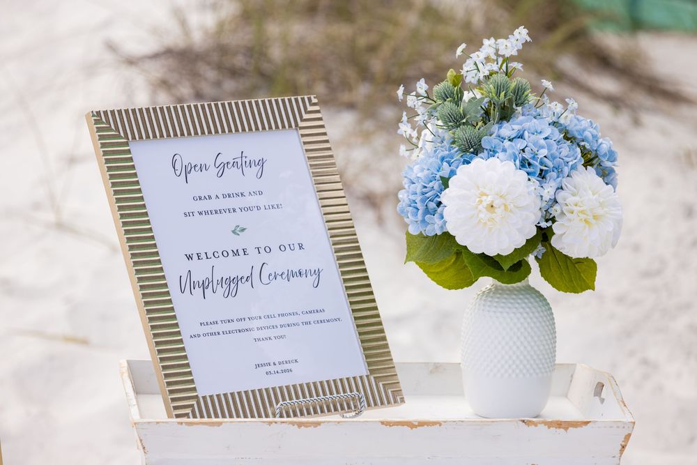 Blue and white hydrangea floral arrangement beside an unplugged wedding ceremony sign on a white tray at a beach wedding.