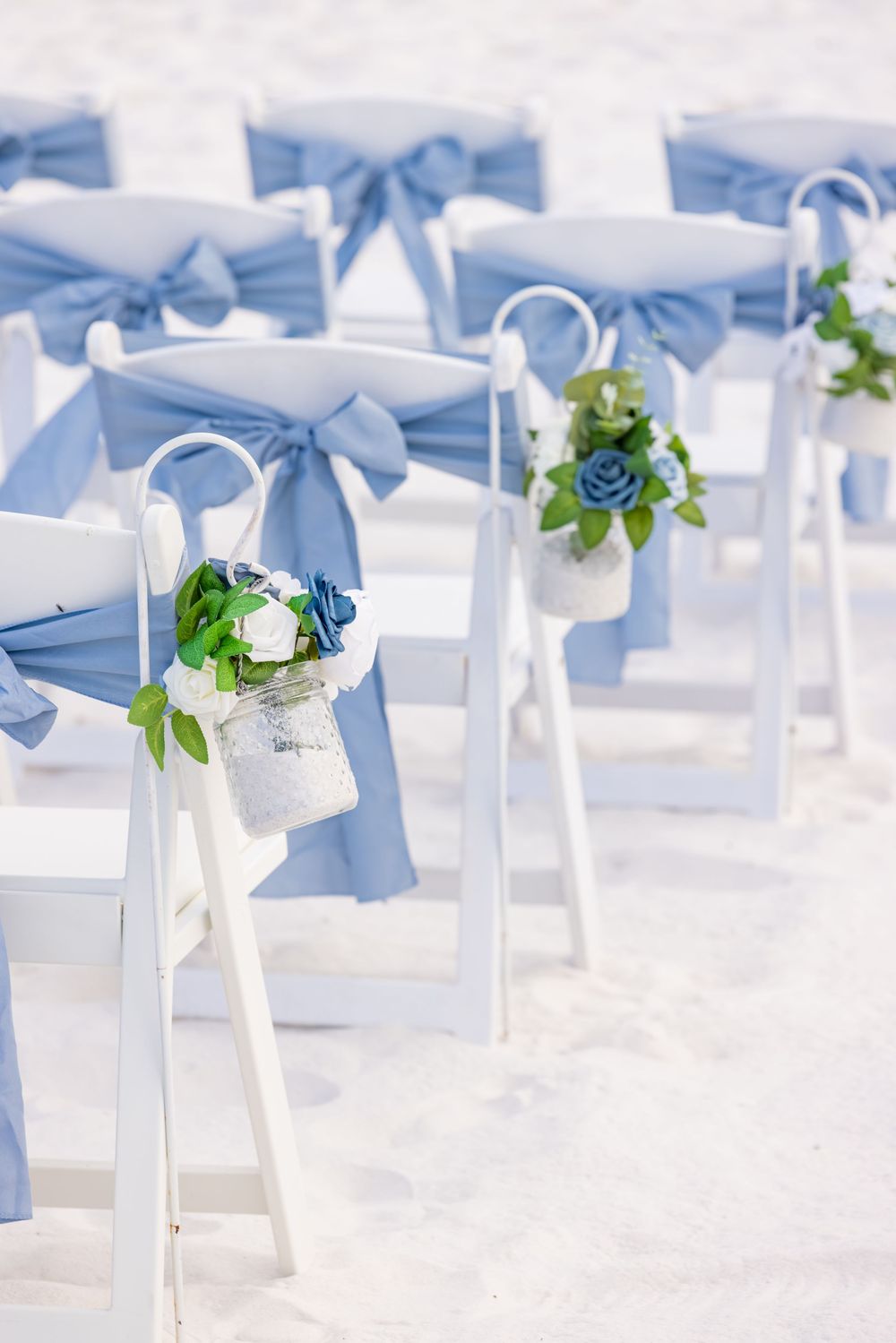 White wedding chairs with blue satin bows and floral arrangements on a sandy beach ceremony setup.