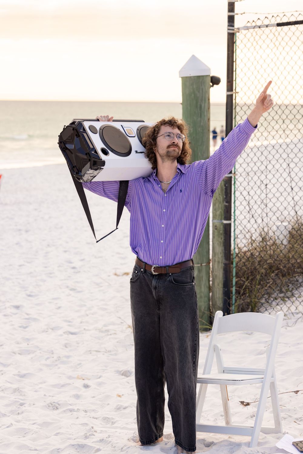 Man holding boombox on shoulder at beach pointing skyward, wearing purple shirt and dark jeans at sunset.