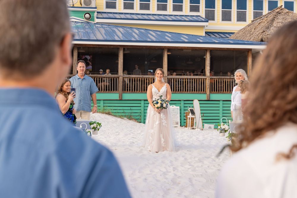 Bride holding bouquet stands on white sand beach during outdoor wedding ceremony with tropical venue backdrop.