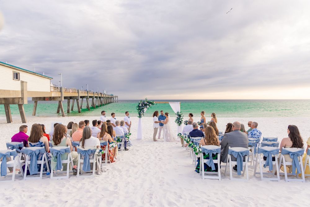 Beach wedding ceremony with guests seated on white chairs facing the ocean near a pier at sunset.
