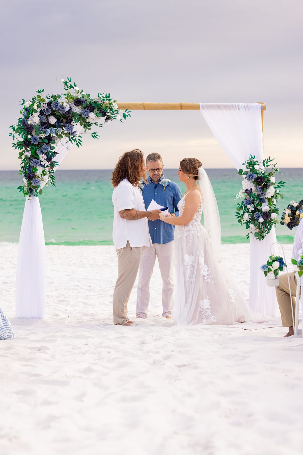 Couple exchanges vows under floral bamboo arch on white sand beach with turquoise ocean backdrop.