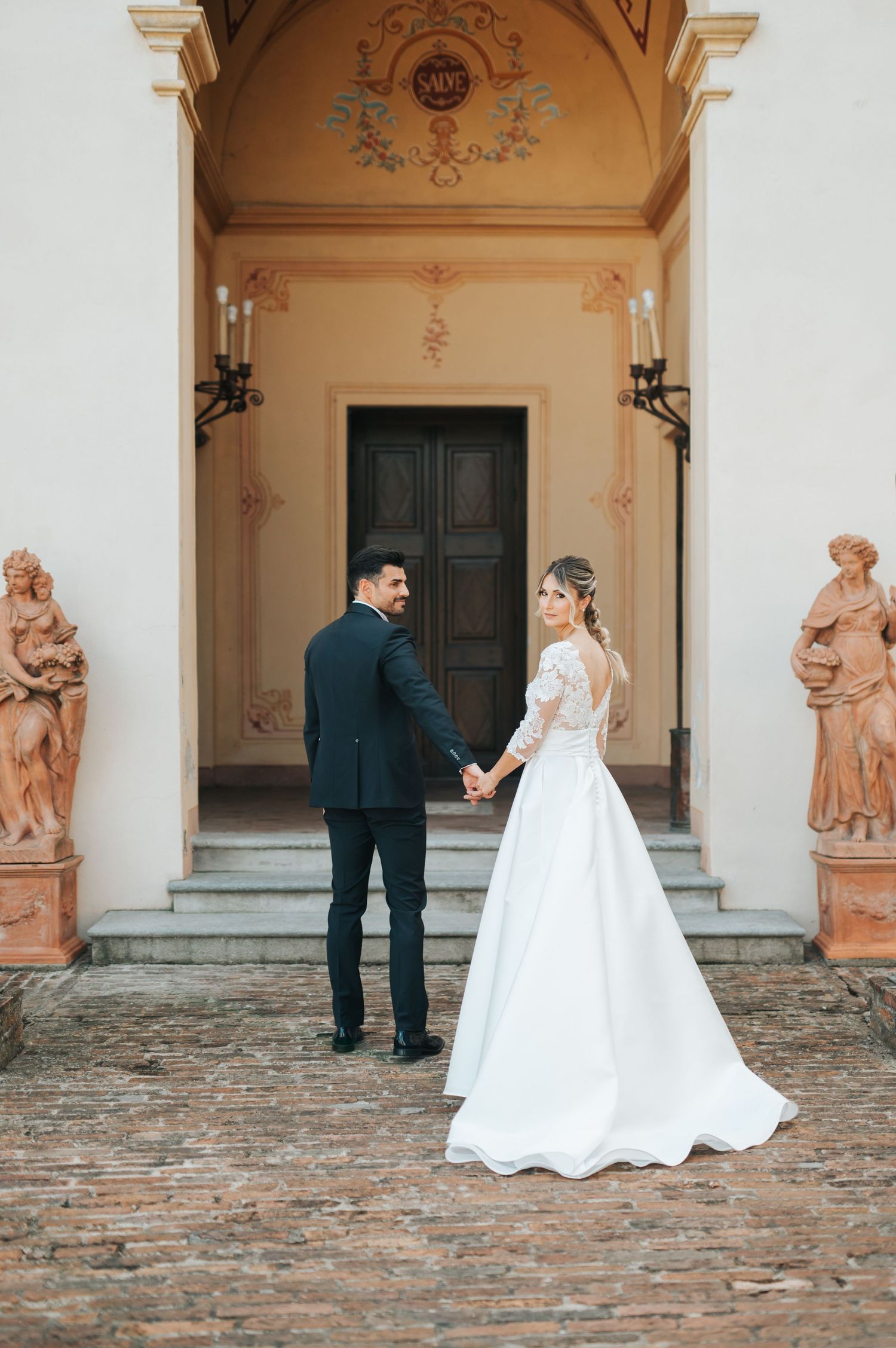 Bride and groom holding hands before ornate church entrance with terracotta statues on wedding day.