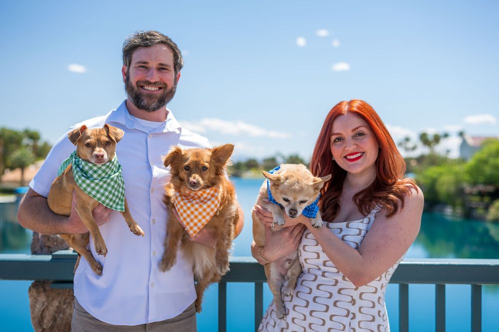 Smiling couple holding three small dogs with bandanas standing by a lake on a sunny day.