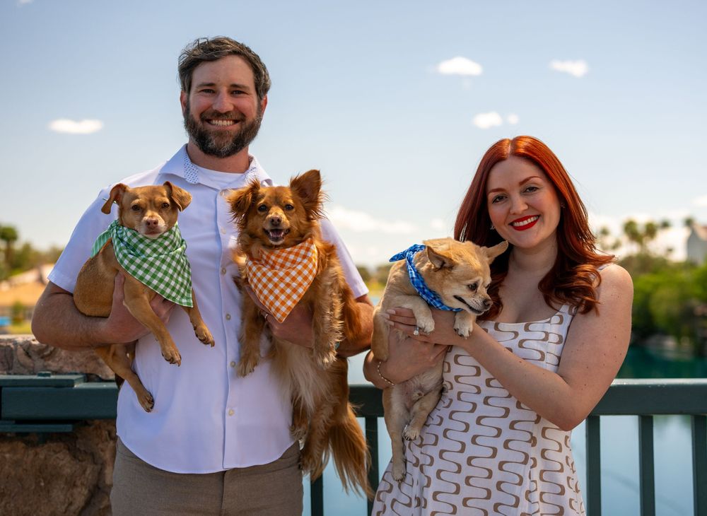 Happy couple holding three small dogs wearing bandanas outdoors near a scenic waterfront at dusk.
