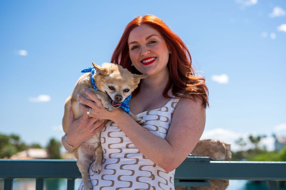 Smiling redhead woman holding a small Chihuahua wearing a blue bow on a sunny day outdoors.
