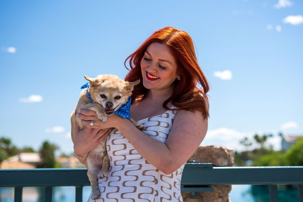 Redhead woman smiling while holding a small Chihuahua with a blue bow outdoors by water.