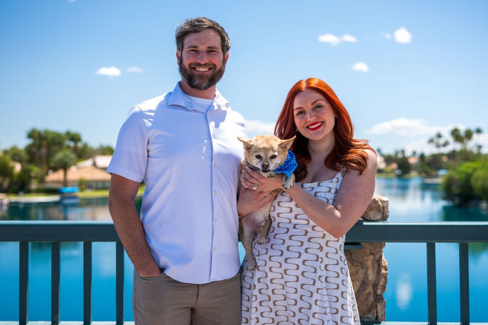 Couple smiling together holding a small Chihuahua on a bridge with a scenic lake and blue sky behind them.