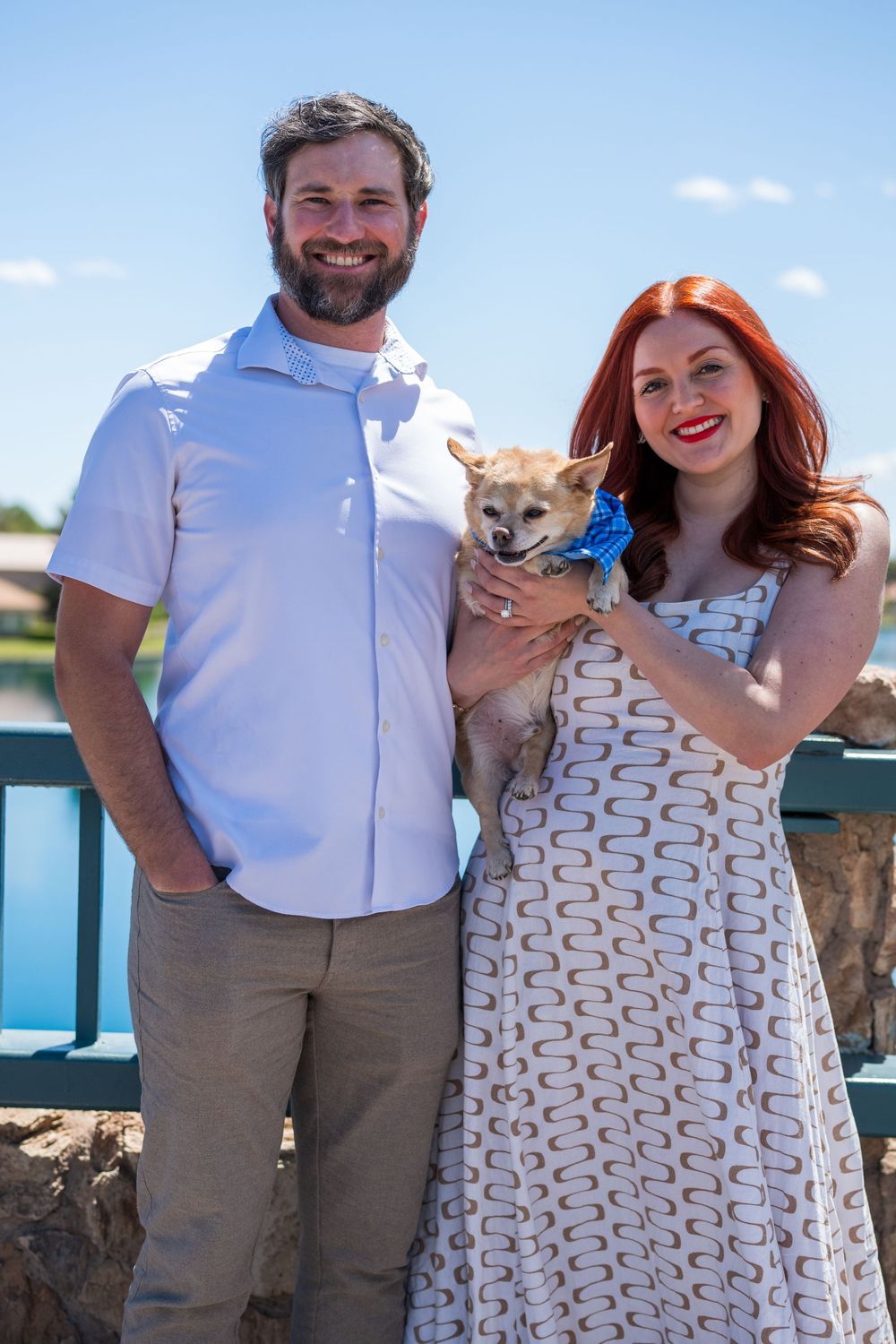 Smiling couple standing outdoors by a lake holding a small Chihuahua with a blue bow.