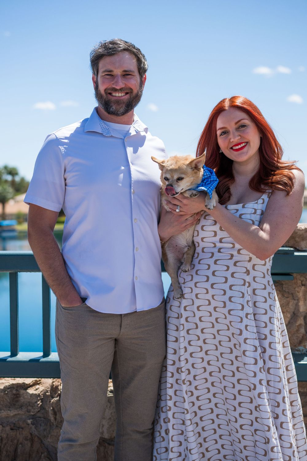 Smiling couple posing outdoors by a lake holding a small Chihuahua wearing a blue bow.