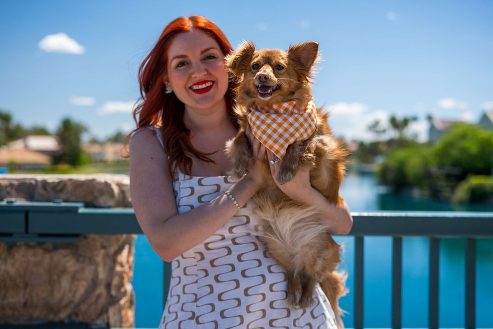 Smiling redhead woman holding a happy Chihuahua in an orange plaid bandana with a bright blue sky backdrop.