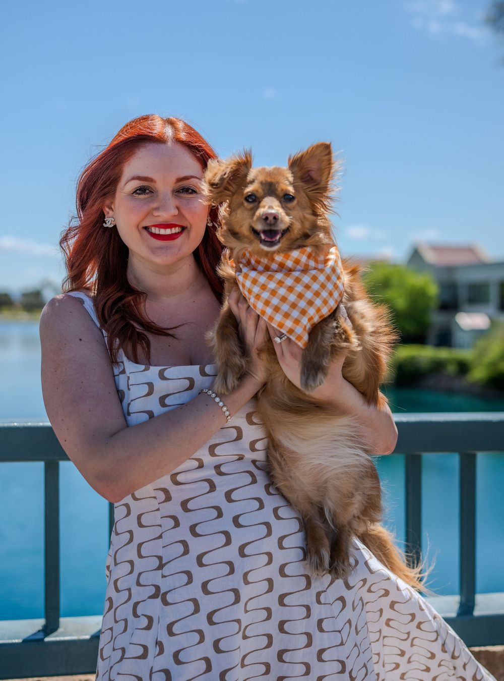Smiling redhead woman holding a Chihuahua in an orange plaid bandana near a scenic waterfront on a sunny day.