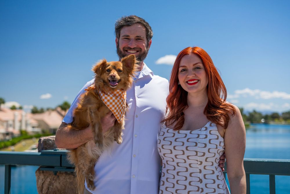 Smiling couple holding a Chihuahua in an orange bandana on a sunny bridge with a blue lake in the background.