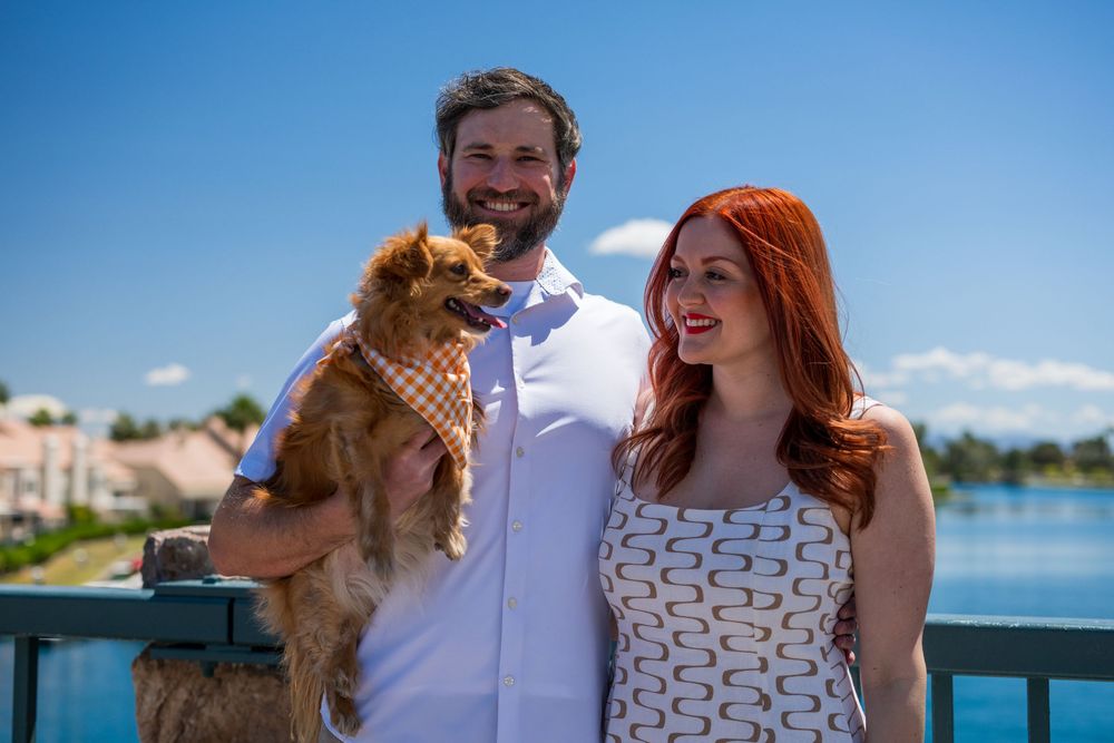 Happy couple with red-haired woman and bearded man holding a small dog by a scenic lake.