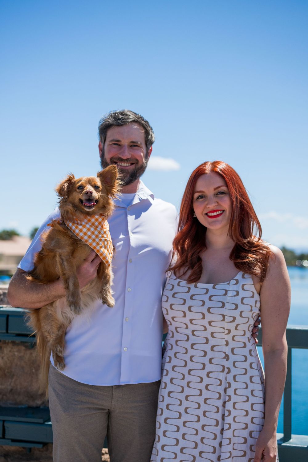 Smiling couple holding a small brown dog wearing a plaid bandana on a sunny waterfront bridge.
