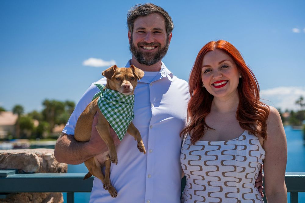 Happy couple holding a small dog wearing a green bandana on a sunny outdoor bridge.