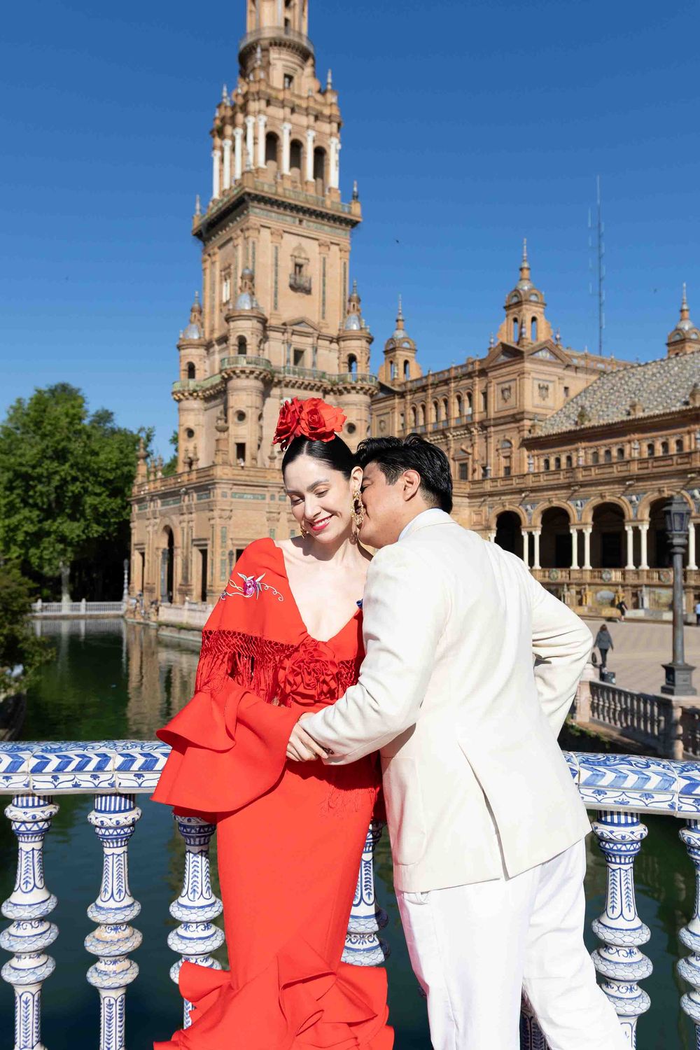 Couple in traditional flamenco attire embracing on a bridge at Plaza de España, Seville, Spain.