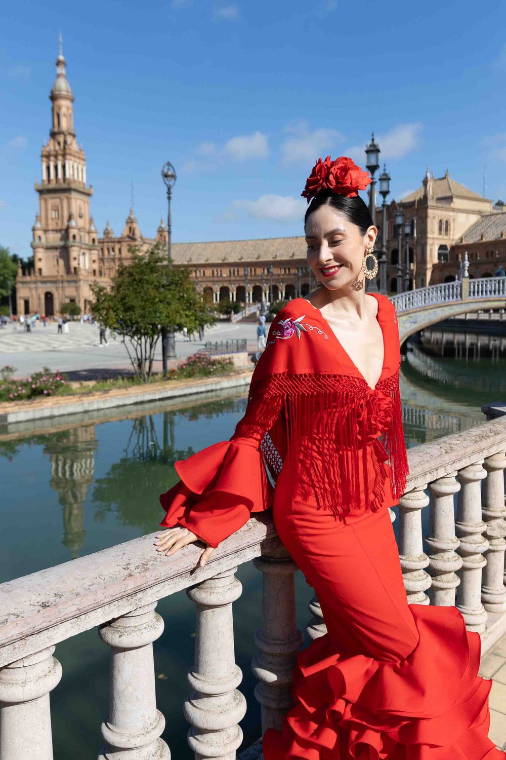 Flamenco dancer in red dress poses at Plaza de España, Seville, with ornate tower and canal in background.