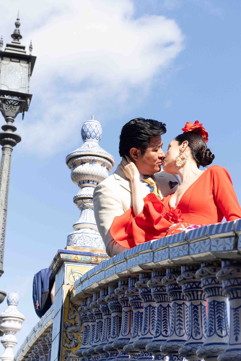 Couple in traditional Spanish flamenco attire sharing a romantic kiss on ornate tiled balcony in Seville, Spain.