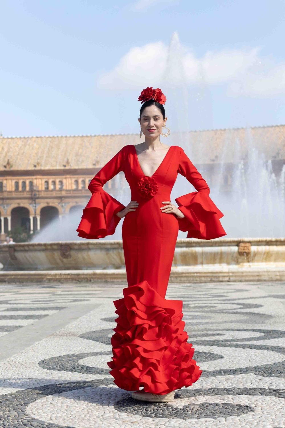 Woman in red flamenco dress with ruffles and flower hair accessory poses at Plaza de España fountain, Seville.