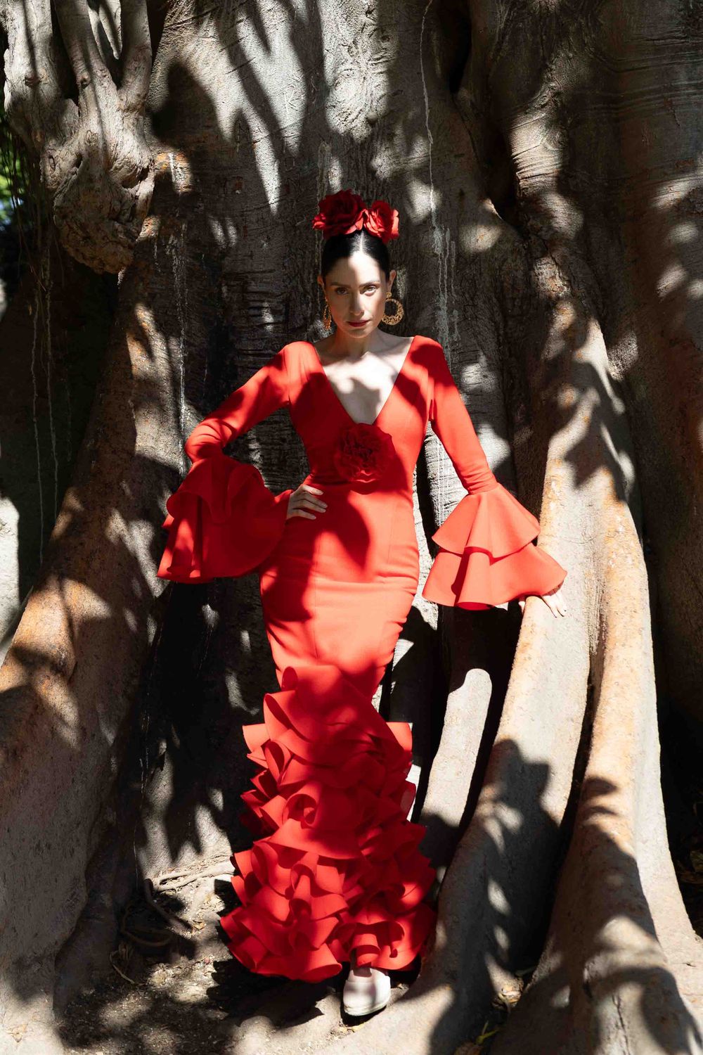 Woman in red flamenco dress with ruffled sleeves poses between large tree trunks in dappled sunlight.