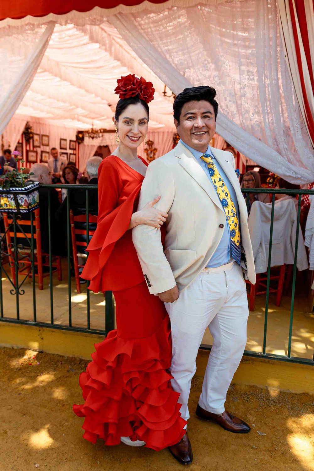 Couple in traditional Spanish flamenco attire posing at a festive venue, woman in red ruffled dress with rose hair accessory.