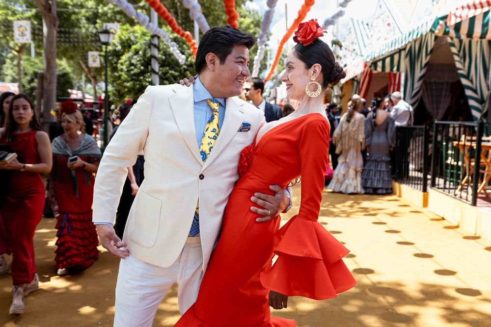 Couple in elegant attire at a Spanish feria, man in white suit, woman in red flamenco dress with flower.