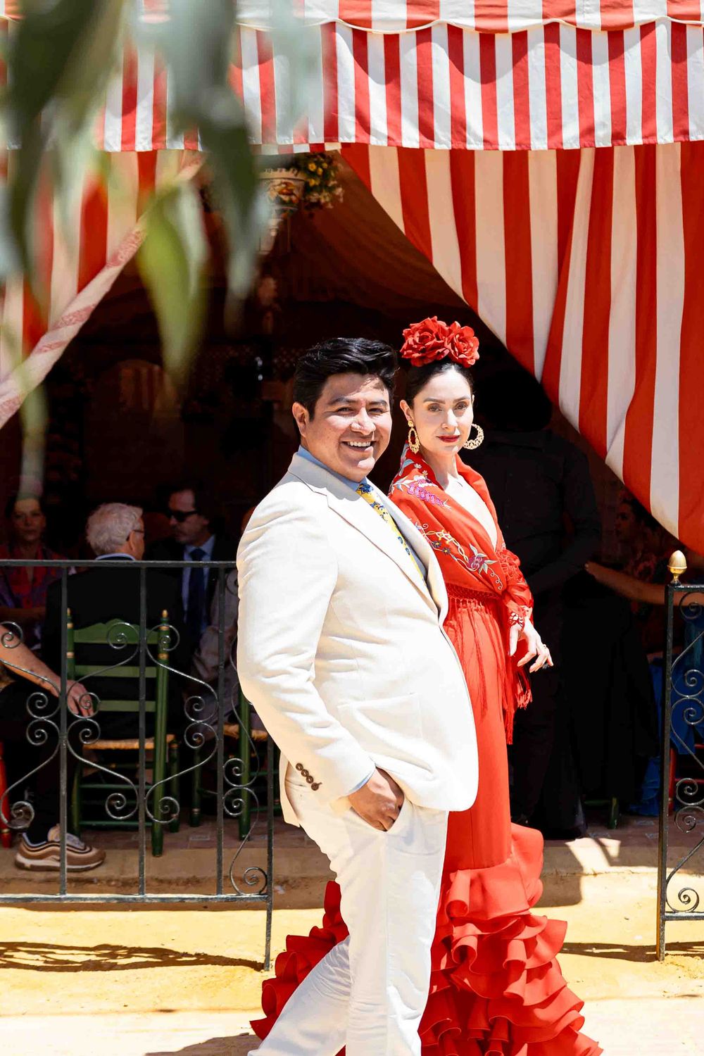 Couple in traditional Spanish flamenco attire posing before a red and white striped tent at Feria de Abril.