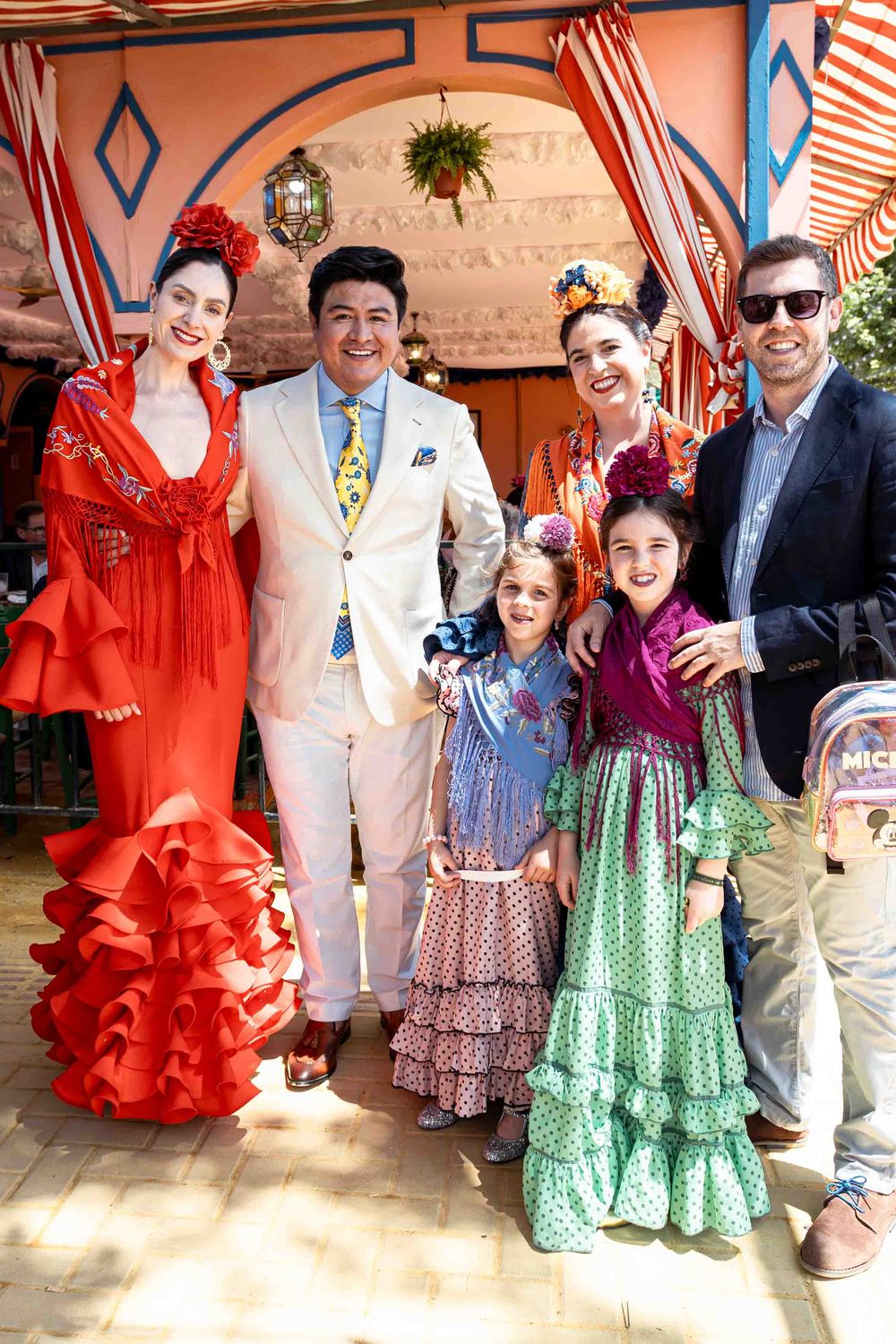 Group of adults and children in vibrant flamenco dresses and festive attire posing together at a colorful Spanish fair.