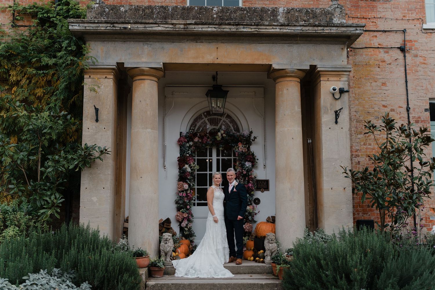 Bride and groom pose on steps of elegant Georgian manor house entrance with columns and autumn floral decorations.