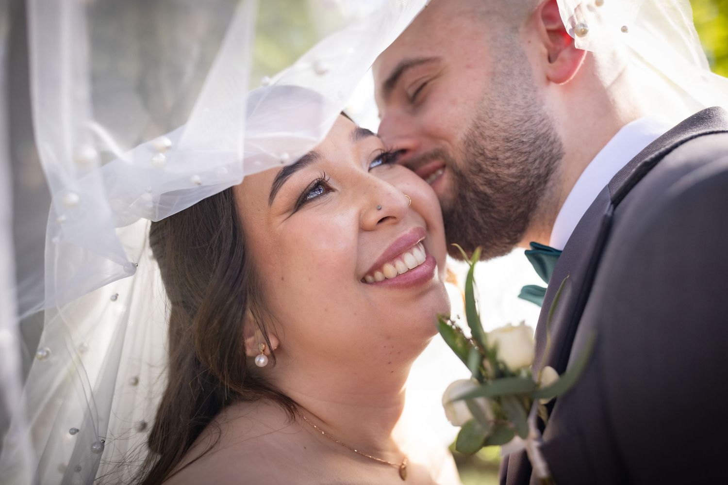 Bride smiling joyfully as groom kisses her cheek beneath a pearl-adorned wedding veil outdoors.