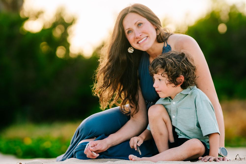 A smiling mother with long dark hair sits outdoors with her young curly-haired son at golden hour.