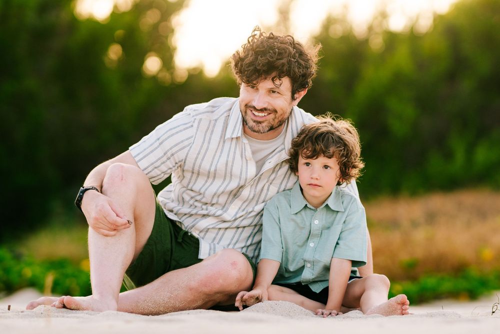 A smiling father crouches beside his young curly-haired son on a sunny sandy beach.