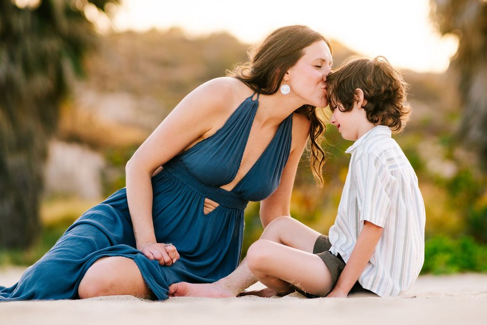 A woman in a blue dress kisses a young boy on the head while sitting on a sandy beach at sunset.