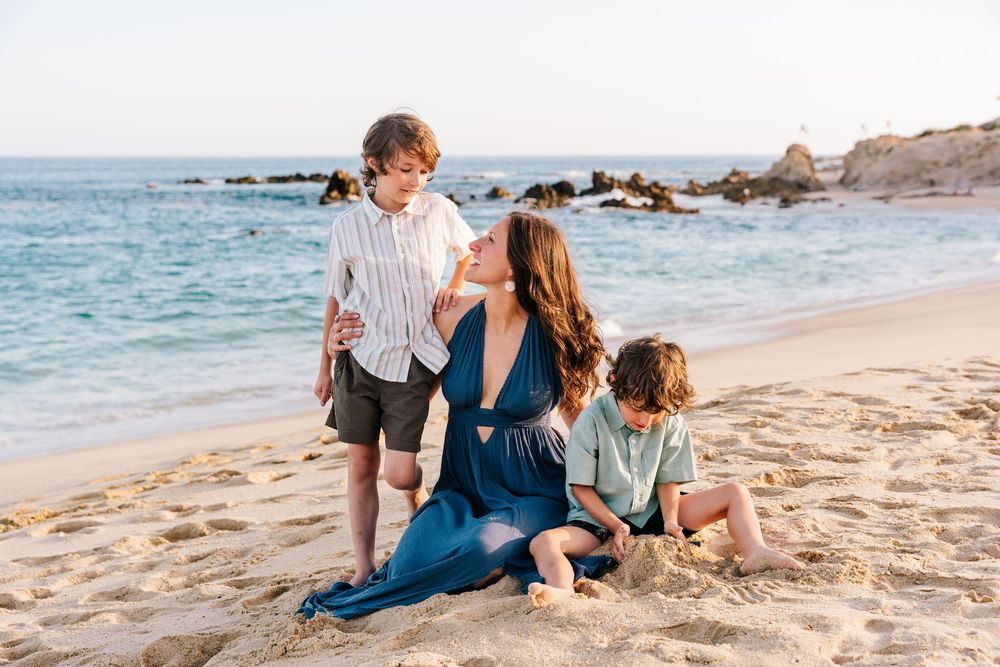 A mother in a blue dress sits on a sandy beach with her two young sons on a sunny day.