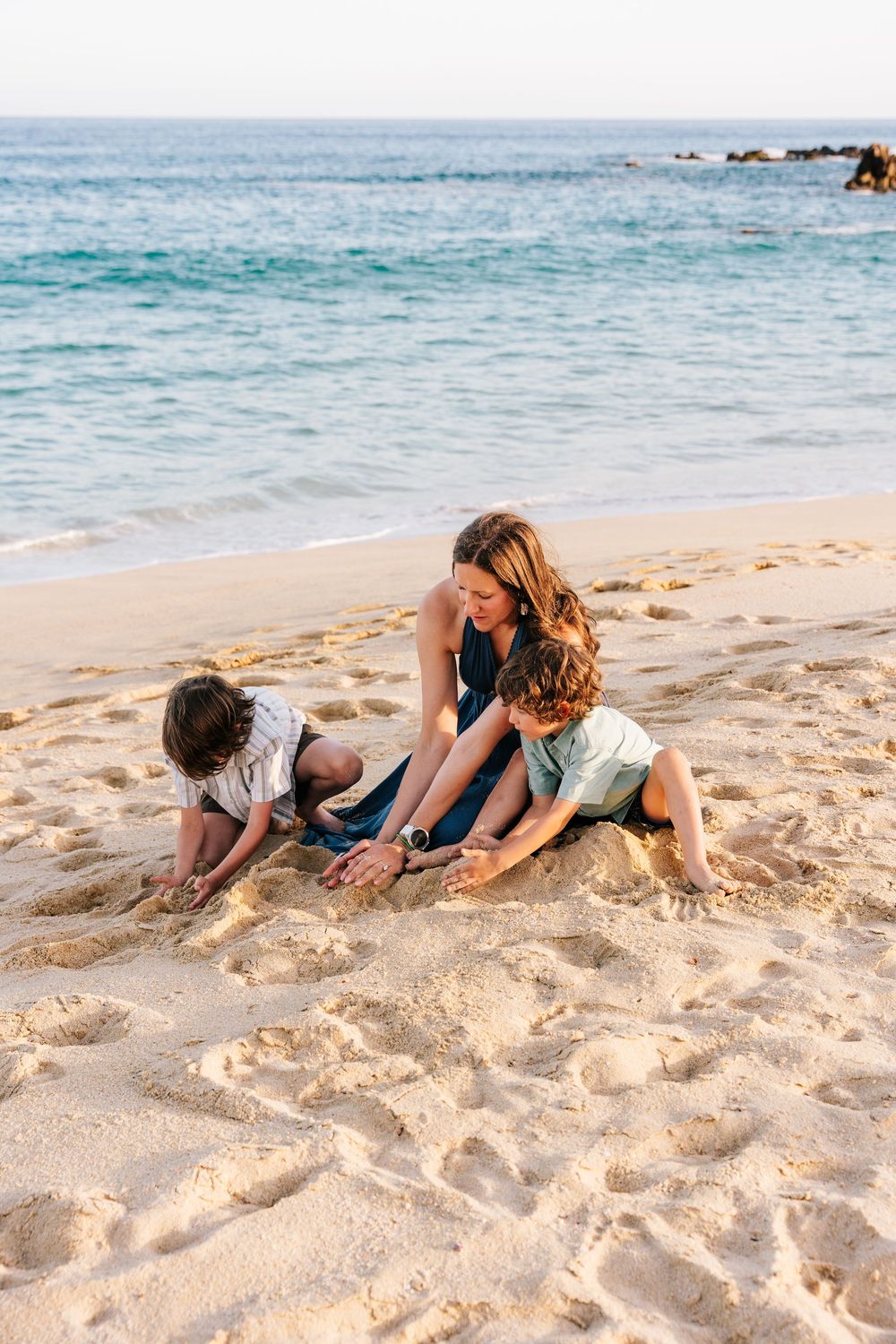 A mother plays in the sand with her two young children on a sunny beach with turquoise ocean waves in the background.