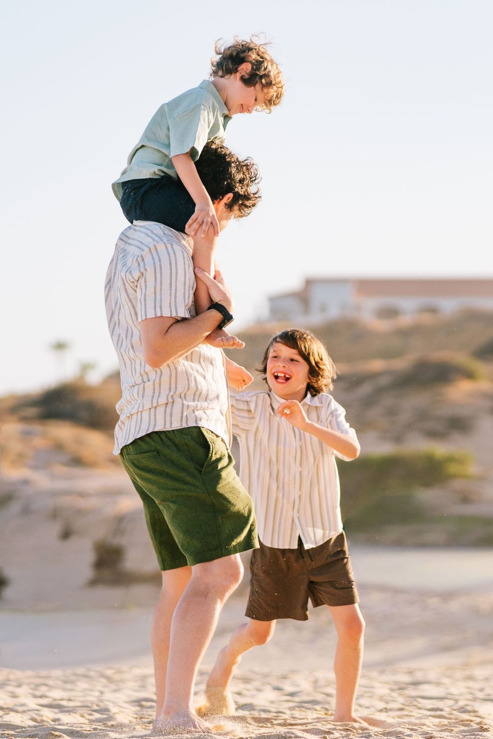 A father carries his son on his shoulders while another child laughs on a sunny beach.