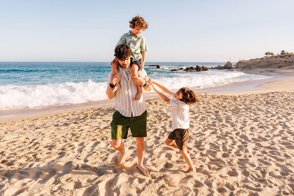 A father plays with his children on a sunny beach, with one child on his shoulders and another dancing nearby.