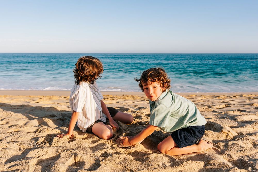 Two curly-haired boys sit together playing in the sand on a sunny beach.
