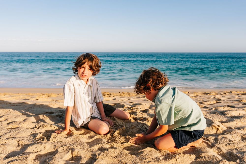 Two young boys play together in the sand on a sunny beach with blue ocean waves behind them.
