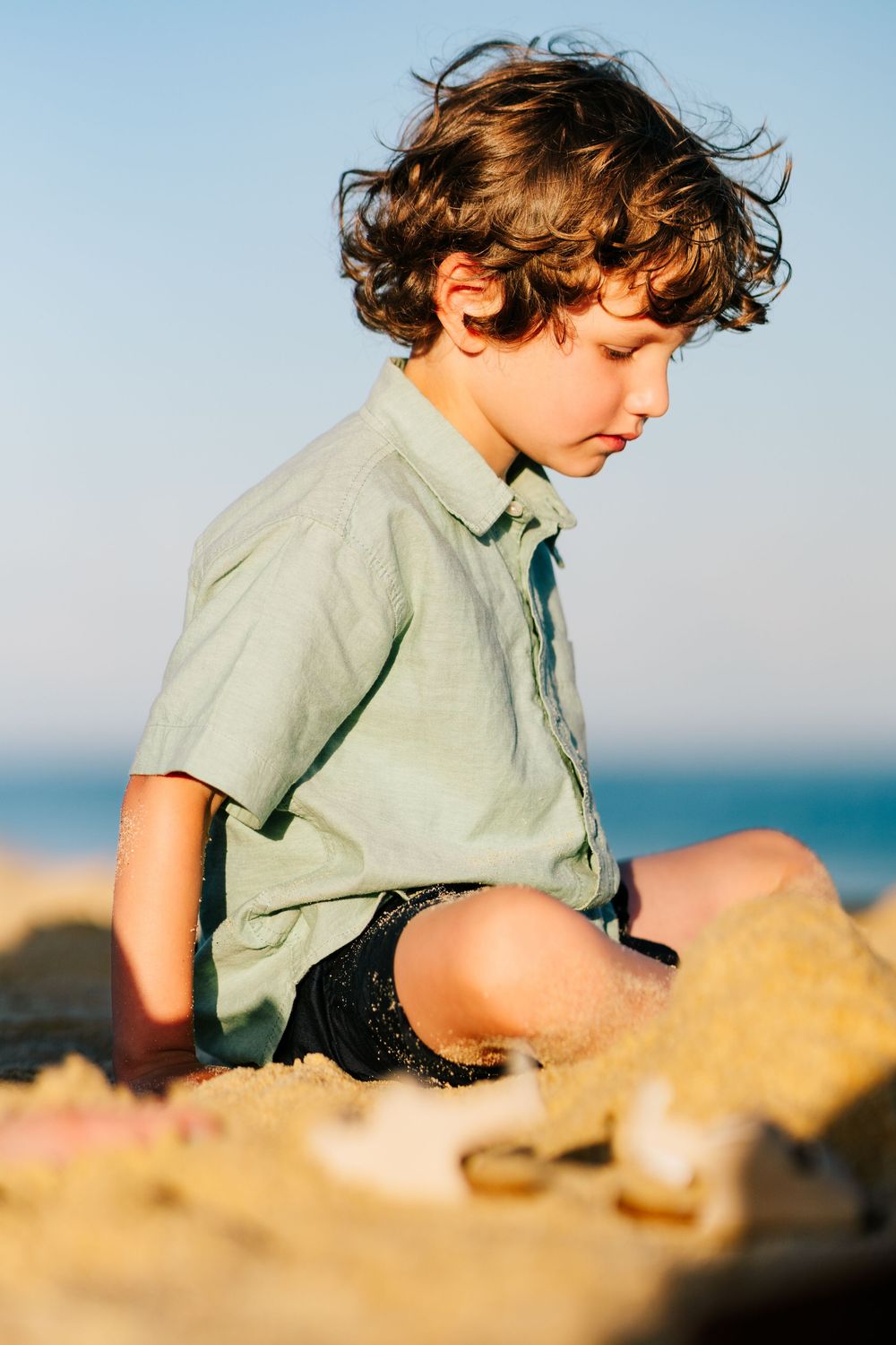 A young curly-haired boy in a green shirt sits quietly playing in the beach sand.