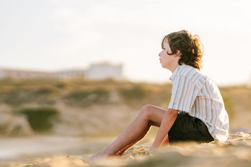 A curly-haired boy sits thoughtfully on sandy dunes at golden hour, gazing into the distance at the beach.