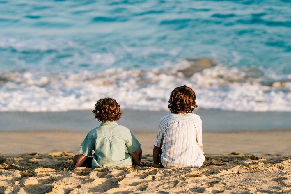 Two young children sitting together on a sandy beach, watching ocean waves from behind.