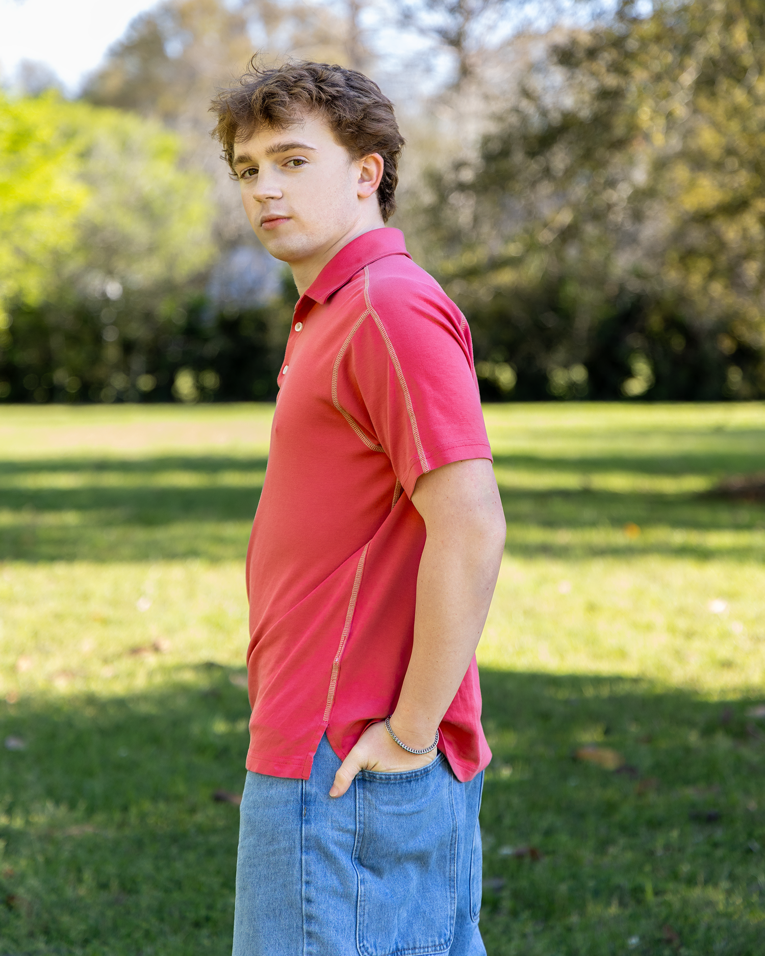 Teen boy in a red polo shirt stands sideways in a grassy park, looking over his shoulder.