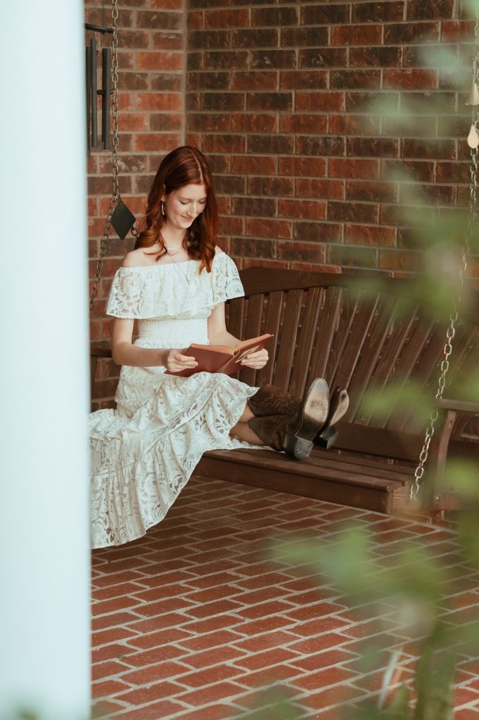 Red-haired woman in white lace off-shoulder dress reading on a wooden porch swing against brick wall.