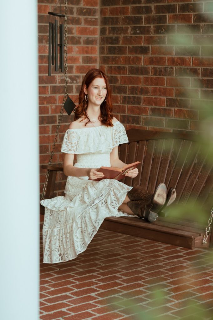 Young woman in white lace off-shoulder dress reading a book on a wooden porch swing against brick wall.