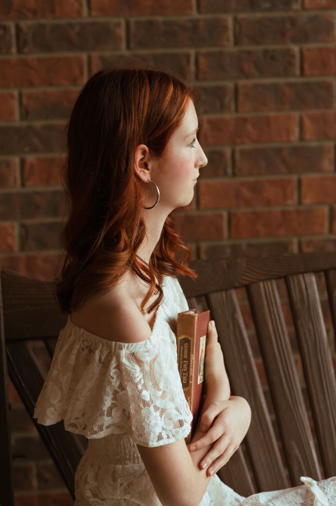 Young redhead woman in white lace off-shoulder dress holding books, seated by brick wall.