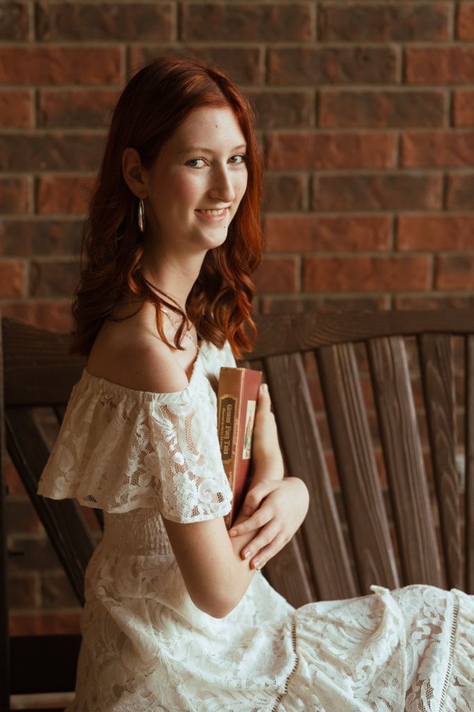 Young woman with red hair in white lace off-shoulder dress holding books, smiling on wooden bench by brick wall.