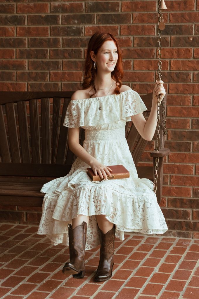 Redhead woman in white lace off-shoulder dress and cowboy boots sitting on a wooden swing holding a book.